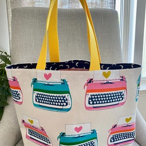 May include: A tote bag with a cream-colored background featuring a repeating pattern of colorful typewriters in shades of teal and pink. Each typewriter has a sheet of paper with a heart icon. The bag includes two bright yellow fabric handles.