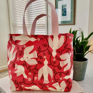 May include: A red tote bag with a white bird and floral pattern. The bag has pink handles and a matching trim. The bag is sitting on a white surface.