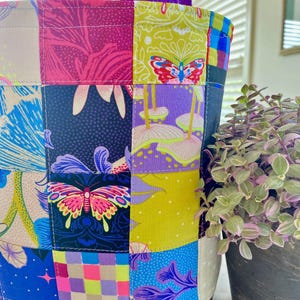 May include: A patchwork tote bag with a purple handle. The bag features a variety of colorful fabric squares with floral and butterfly patterns. The bag's base is a dark maroon fabric, and the handle is a solid purple.