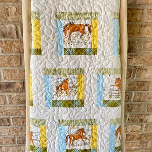 May include: A rectangular quilt featuring a repeating pattern of brown horses in a landscape, framed by yellow, light blue, and green plaid fabric borders. The white background fabric has a continuous swirling quilted texture throughout the surface.
