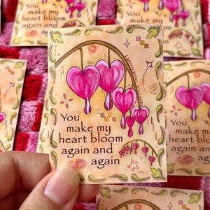 May include: A collection of cards featuring a watercolor illustration of pink bleeding hearts. The cards have the text "You make my heart bloom again and again" in a decorative font. The background is a soft beige with floral accents.