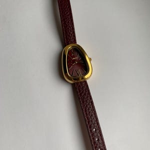 May include: A gold-tone watch with a unique, asymmetrical, teardrop-shaped face. The watch has a burgundy leather band.