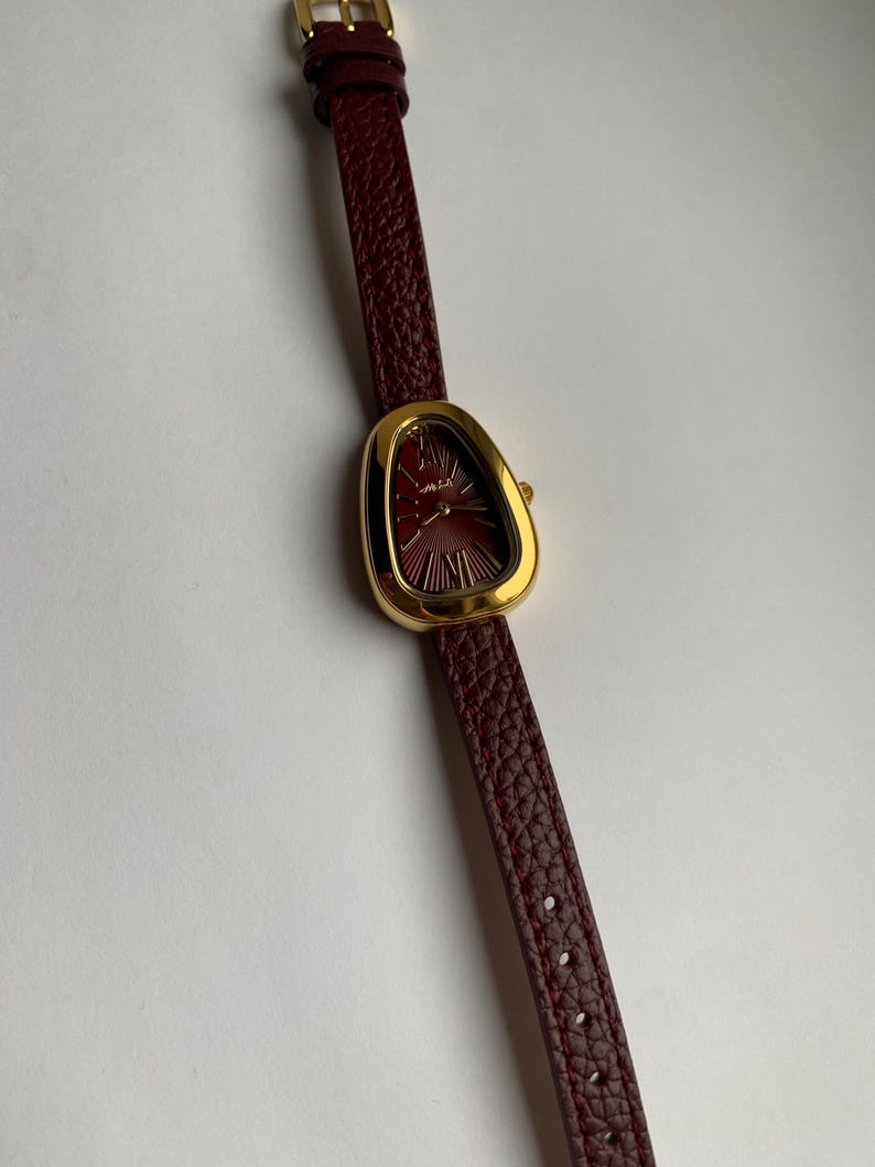 May include: A gold-tone watch with a unique, asymmetrical, teardrop-shaped face. The watch has a burgundy leather band.