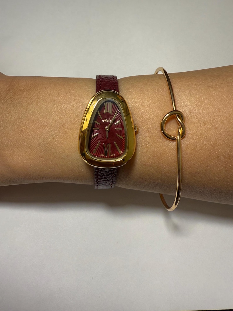 May include: A gold-tone watch with a red face and Roman numeral hour markers. The watch has a burgundy leather band and is worn on a person's wrist. A gold bangle bracelet with a knot detail is also worn on the wrist.