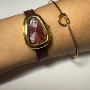 May include: A gold-tone watch with a burgundy leather band and a unique, asymmetrical, oval-shaped face. The watch is worn on a wrist with a gold bangle bracelet.