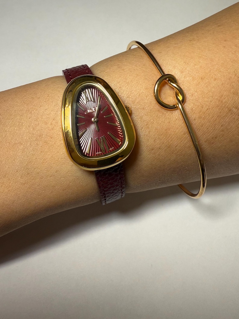 May include: A gold-tone watch with a burgundy leather band and a unique, asymmetrical, oval-shaped face. The watch is worn on a wrist with a gold bangle bracelet.