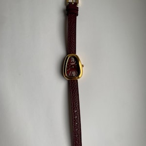 May include: A gold-tone watch with a burgundy leather band. The watch face is oval shaped and has a red dial.