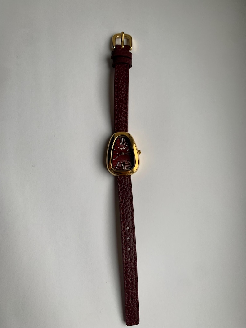 May include: A gold-tone watch with a burgundy leather band. The watch face is oval shaped and has a red dial.