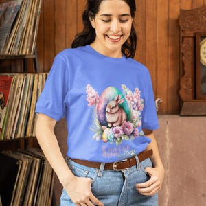 Puede incluir: Una mujer lleva una camiseta azul claro con una ilustración de acuarela de un conejo dentro de un huevo pastel rodeado de flores. El texto "Easter Vibes" está impreso debajo de la ilustración. Lleva pantalones vaqueros azules y un cinturón marrón.