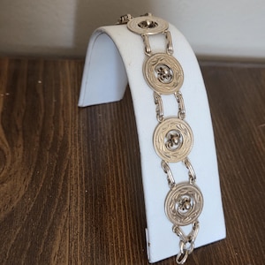 May include: A silver-toned bracelet with circular links, each featuring a central floral design and a decorative border. The bracelet is displayed on a white stand against a wooden surface.