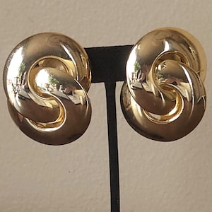 May include: A pair of gold-toned clip-on earrings. Each earring features a circular design with an interlocking, figure-eight shape. The earrings have a polished, reflective surface, and are displayed on a black stand against a neutral background.
