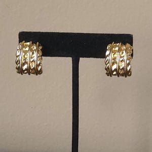 May include: A pair of gold-colored hoop earrings with a textured, braided design. The earrings are displayed on a black stand against a neutral background. The earrings have a classic, elegant style.