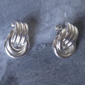 May include: A pair of polished silver-tone clip-on earrings featuring a multi-layered, interlocking hoop design. Each earring consists of three smooth, rounded metal bands that overlap to create a dimensional, circular shape with a secure screw-back mechanism.