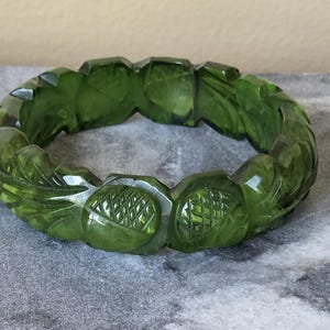 Puede incluir: Un brazalete verde con diseños tallados de hojas y flores. El brazalete está hecho de un material translúcido verde oliva y descansa sobre una superficie gris. Es una pieza de joyería.