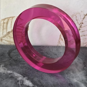 May include: A circular bangle bracelet made of translucent, vibrant fuchsia pink acrylic material. The object features a smooth, polished surface with a thick, rounded profile and a hollow center, showcasing a clear and glossy finish throughout.