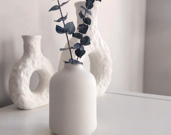 May include: A white ceramic vase with a textured surface holds a sprig of dried eucalyptus with blue-grey leaves. The vase is sitting on a white surface.