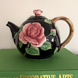 May include: Black ceramic teapot with a large pink rose and green leaves. The handle is brown and the lid is topped with a small rose bud. The teapot is sitting on a green book.