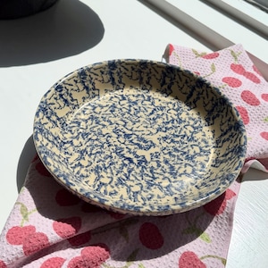 May include: A round ceramic pie dish with a cream-colored base and a blue splatter pattern. The dish sits on a pink cherry-patterned kitchen towel. The dish is approximately 9 inches in diameter.
