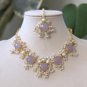 Puede incluir: Un conjunto de joyas de tres piezas, que incluye un collar, una pieza para la cabeza y pendientes. El collar y los pendientes presentan piedras de color lavanda, pequeñas perlas blancas y metal dorado. La pieza para la cabeza tiene un diseño similar.