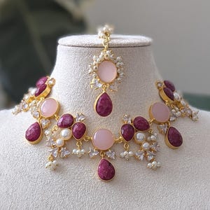 Op de afbeelding: Een goudkleurige sieradenset op een mannequin. De set bevat een ketting, oorbellen en een hoofdband, allemaal versierd met roze en robijnkleurige stenen en kleine witte parels. De ketting en hoofdband hebben een cirkelvormig ontwerp.