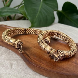 May include: Two gold-colored bangles with intricate floral and geometric patterns. The bangles are displayed on a wooden tray, showcasing their detailed design and craftsmanship. The bangles have a clasp closure.