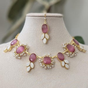 May include: A jewellery set comprising a necklace, earrings, and a headpiece, displayed on a mannequin. The set features pink oval stones, white leaf-shaped accents, and small pearls, all set in gold-coloured metal.