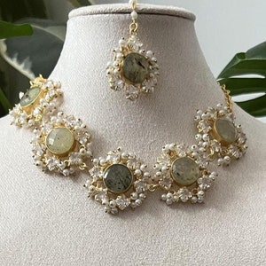May include: A three-piece jewelry set on a mannequin. The set includes a necklace, earrings, and a headpiece. The necklace and earrings feature round, light green stones surrounded by small white pearls and gold-colored metal. The headpiece has a similar design.