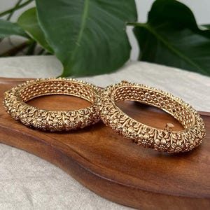 May include: Two gold-colored bangles with intricate floral patterns. The bangles are displayed on a wooden surface, showcasing their detailed design and texture. The jewelry is a classic accessory.
