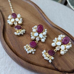 May include: A gold-coloured jewellery set with ruby-coloured gemstones, white stones, and small pearls. The set includes a headpiece and two pairs of earrings, arranged on a wooden tray.