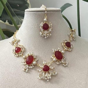 May include: A gold-coloured necklace and headpiece set. The necklace features large, oval, and triangular red gemstones surrounded by small white pearls. The headpiece has a matching red gemstone and pearl accents. The jewellery is displayed on a mannequin.