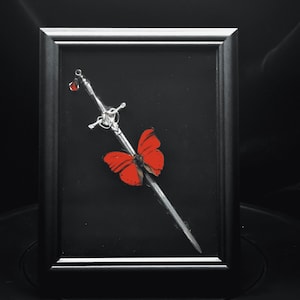 May include: A framed display featuring a silver-toned sword positioned diagonally against a black background. A vibrant red butterfly is attached to the blade, and a small, translucent red teardrop-shaped charm hangs from the sword's hilt.