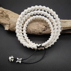 May include: A multi-layered bracelet constructed from white, segmented vertebrae bones arranged in a circular pattern. The structure is held together by a thin black cord that extends into two loose ends, each finished with a single small bone bead.
