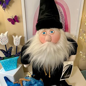 May include: A plush wizard doll with a long white beard, blue eyes, and a black hat and robe. A blue dragon plush with gold accents sits in front of the wizard. Decorative tulips and butterflies are in the background.