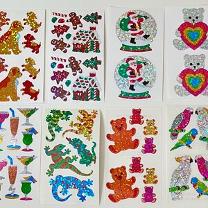 May include: Eight sheets of holographic stickers featuring various designs including golden dogs, gingerbread houses, Santa Claus in snow globes, teddy bears with rainbow hearts, colorful cocktails, lizards, gummy bears, and tropical parrots.