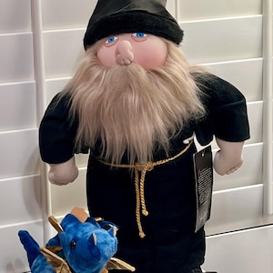 May include: A plush wizard doll wearing a black robe and matching pointed hat with a long beige beard and blue eyes, accompanied by a small blue plush dragon with gold wings and a tan belly. A black product tag hangs from the wizard's waist.