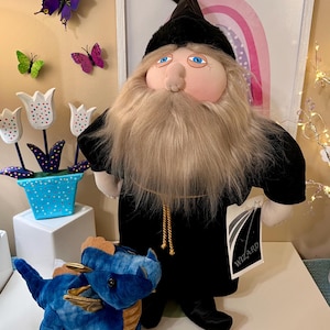 May include: A plush wizard doll with a long, beige beard and a black robe, standing next to a blue stuffed dragon with gold accents. The wizard has a black hat and a tag that says "WIZARD". Decorative butterflies and tulips are in the background.