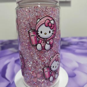 May include: A clear glass jar filled with pink glitter and decorated with Hello Kitty stickers. The stickers show Hello Kitty in a pink hoodie holding a pink Stanley cup. The jar has a white lid.