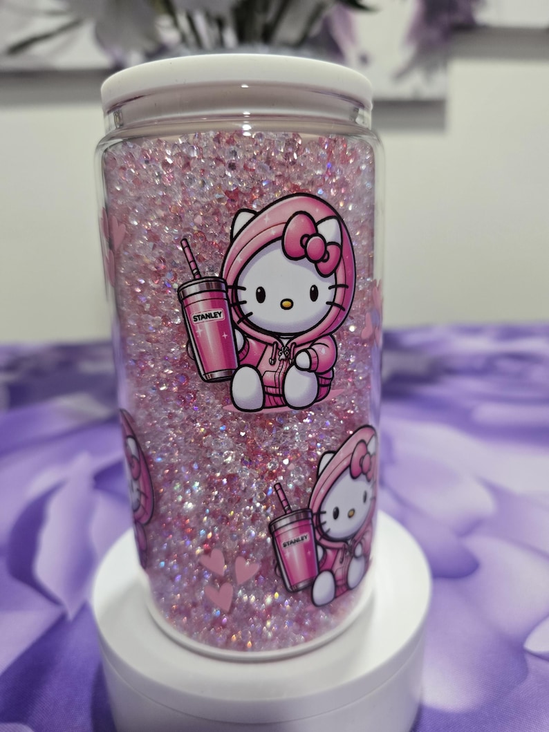 May include: A clear glass jar filled with pink glitter and decorated with Hello Kitty stickers. The stickers show Hello Kitty in a pink hoodie holding a pink Stanley cup. The jar has a white lid.