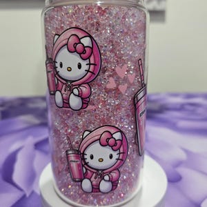 May include: A clear tumbler filled with pink glitter and featuring Hello Kitty in a pink hoodie. The tumbler has a white lid and is decorated with pink hearts and a pink Stanley cup.