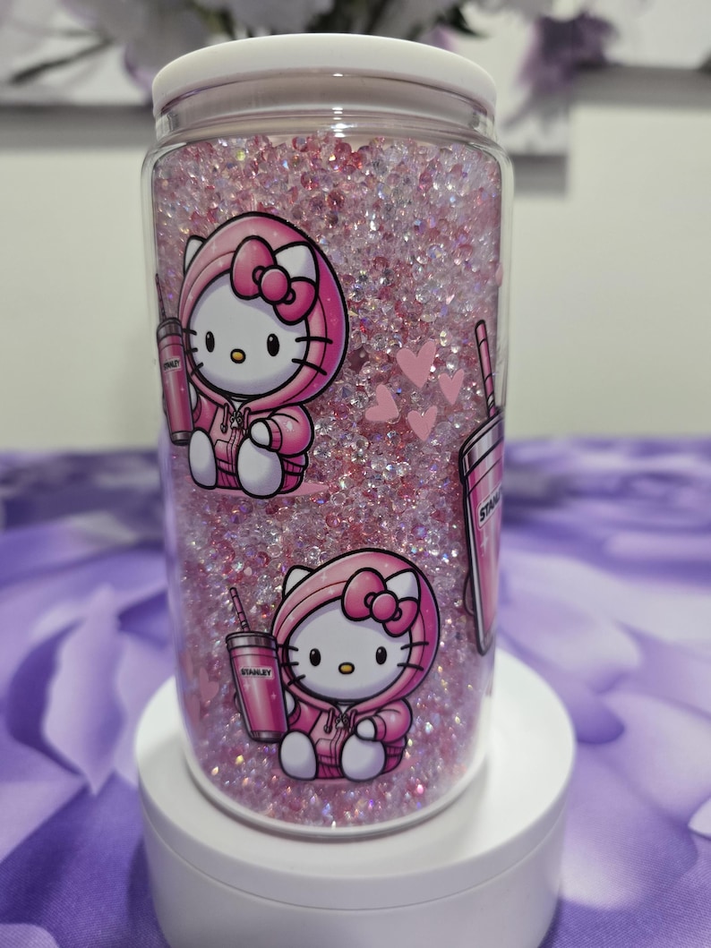 May include: A clear tumbler filled with pink glitter and featuring Hello Kitty in a pink hoodie. The tumbler has a white lid and is decorated with pink hearts and a pink Stanley cup.