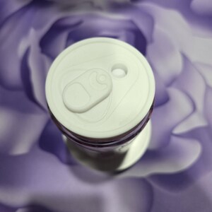 May include: A white aluminum can with a pull tab, viewed from above. The can's top is white, and the body is partially visible with a purple hue. The background is a blurred, abstract pattern of purple and white.