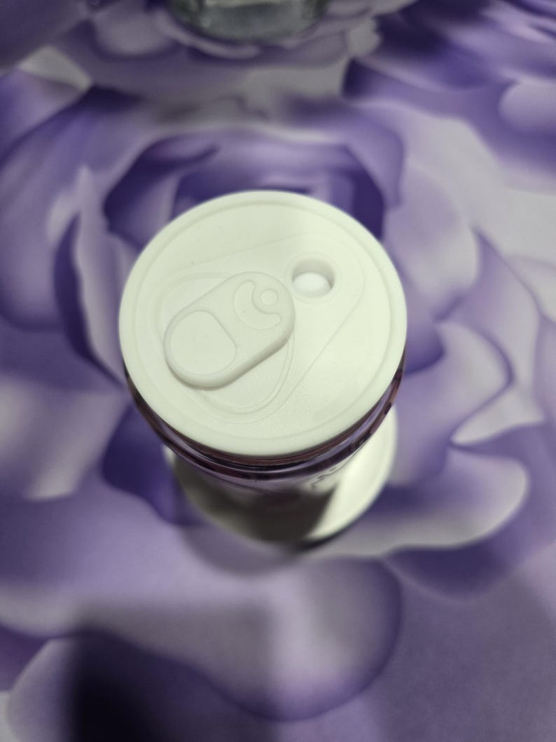 May include: A white aluminum can with a pull tab, viewed from above. The can's top is white, and the body is partially visible with a purple hue. The background is a blurred, abstract pattern of purple and white.
