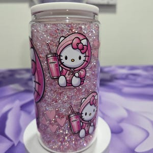 May include: A clear glass tumbler filled with pink glitter and featuring a Hello Kitty design. The design includes Hello Kitty in a pink hoodie holding a pink Stanley tumbler. Pink hearts are also visible.