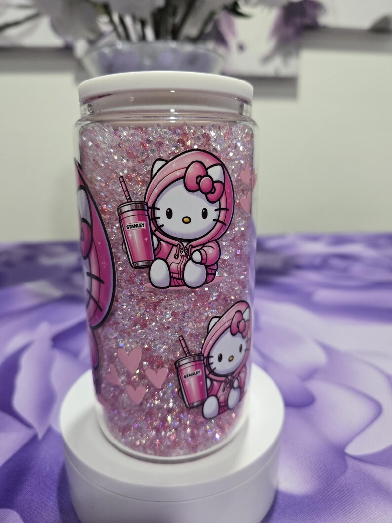 May include: A clear glass tumbler filled with pink glitter and featuring a Hello Kitty design. The design includes Hello Kitty in a pink hoodie holding a pink Stanley tumbler. Pink hearts are also visible.