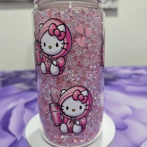 May include: A clear, cylindrical tumbler filled with pink glitter and a white lid. The tumbler features a cartoon image of Hello Kitty wearing a pink hoodie and holding a pink Stanley tumbler. Two pink hearts are also visible.