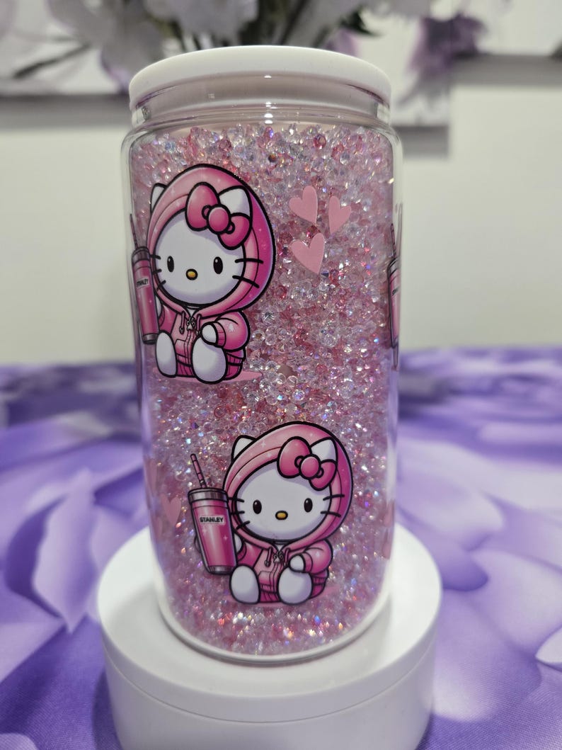 May include: A clear, cylindrical tumbler filled with pink glitter and a white lid. The tumbler features a cartoon image of Hello Kitty wearing a pink hoodie and holding a pink Stanley tumbler. Two pink hearts are also visible.