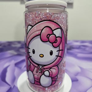 May include: A clear, cylindrical container with a white lid, filled with pink glitter and featuring a Hello Kitty design in a pink hoodie. The container is sitting on a white pedestal.