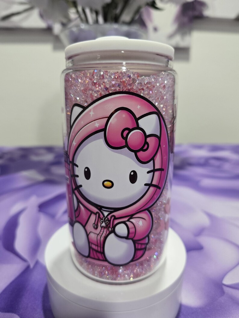 May include: A clear, cylindrical container with a white lid, filled with pink glitter and featuring a Hello Kitty design in a pink hoodie. The container is sitting on a white pedestal.