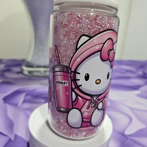 May include: A clear glass tumbler filled with pink glitter and a pink lid. The tumbler features a cartoon graphic of Hello Kitty in a pink hoodie holding a pink Stanley tumbler. The tumbler is on a white stand.