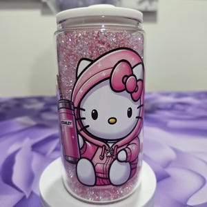 May include: A clear, cylindrical tumbler filled with pink glitter and a cartoon graphic of Hello Kitty in a pink hoodie. A pink Stanley tumbler graphic is also visible. The tumbler has a white lid and sits on a white stand.